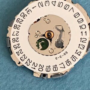 May include: Close-up of a watch movement with a white date wheel featuring black numerals from 1 to 31. The movement has a silver-colored metal frame and various internal components. The text "E6-3-3" is printed on the movement.