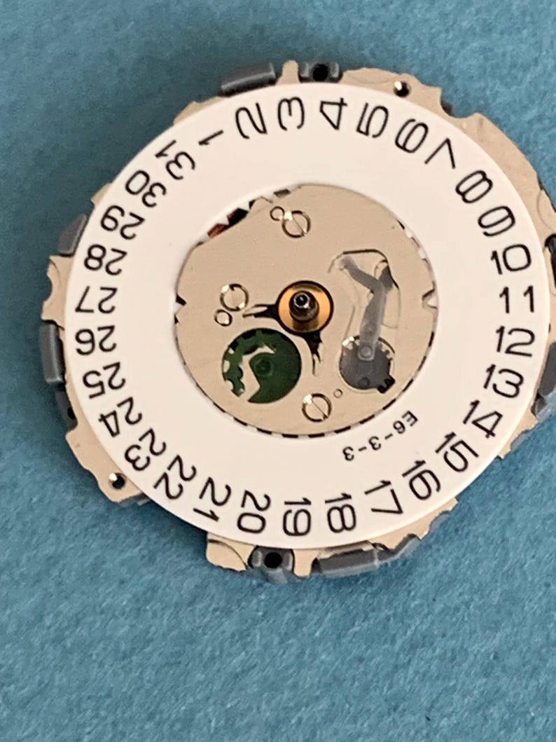 May include: Close-up of a watch movement with a white date wheel featuring black numerals from 1 to 31. The movement has a silver-colored metal frame and various internal components. The text "E6-3-3" is printed on the movement.