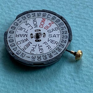K&ouml;nnte beinhalten: Ein Uhrwerk mit wei&szlig;em Zifferblatt, das die Wochentage auf Englisch und die Zahlen 1-31 anzeigt. Der Wochentag ist DOM (Sonntag) in Rot. Eine kleine goldfarbene Schraube ist an der Seite befestigt.