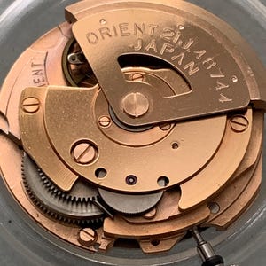 Puede incluir: Primer plano de un mecanismo de reloj con engranajes y componentes dorados. El texto "ORIENT 21J JAPAN 46744" es visible en una placa curva. El diseño intrincado muestra la mecánica interna de un reloj.