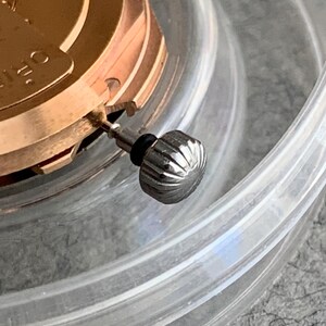 Puede incluir: Primer plano de un movimiento de reloj con una corona plateada. La corona es estriada y tiene una junta de goma negra. El mecanismo del reloj es de color dorado y parcialmente visible dentro de un recipiente de pl&aacute;stico transparente.