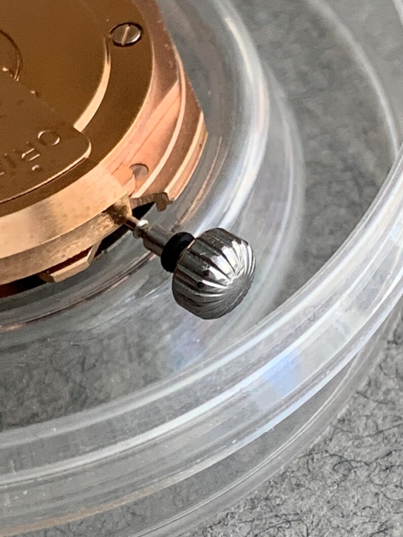 Puede incluir: Primer plano de un movimiento de reloj con una corona plateada. La corona es estriada y tiene una junta de goma negra. El mecanismo del reloj es de color dorado y parcialmente visible dentro de un recipiente de pl&aacute;stico transparente.