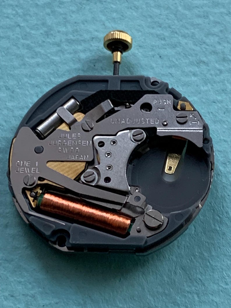 K&ouml;nnte beinhalten: Nahaufnahme eines Jules Jurgensen Uhrwerks. Der runde Mechanismus weist verschiedene Metallkomponenten auf, darunter eine Kupferspule und eine goldfarbene Krone. Der Text "Jules Jurgensen SW10 Japan" ist sichtbar. Der Hintergrund ist hellblau.