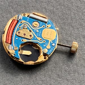 May include: Close-up of a watch movement with a blue circuit board, gold-colored metal components, and a small screw. The watch movement has the text "ZENITH" and "SWISS V8" printed on it. The watch movement is a small, intricate piece of machinery.