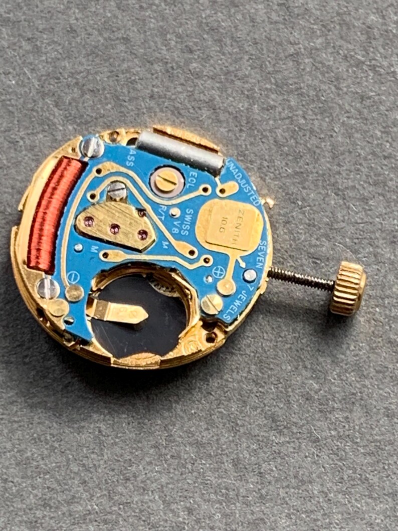 May include: Close-up of a watch movement with a blue circuit board, gold-colored metal components, and a small screw. The watch movement has the text "ZENITH" and "SWISS V8" printed on it. The watch movement is a small, intricate piece of machinery.