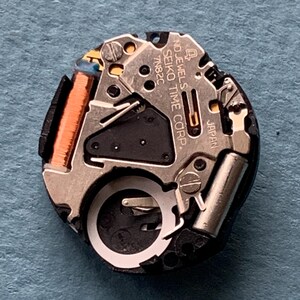 K&ouml;nnte beinhalten: Nahaufnahme eines Seiko-Uhrwerks. Der Mechanismus umfasst silberfarbene Metallkomponenten, eine kupferfarbene Spule und den Text "SEIKO TIME CORP" und "JAPAN". Das Uhrwerk befindet sich vor einem blauen Hintergrund.