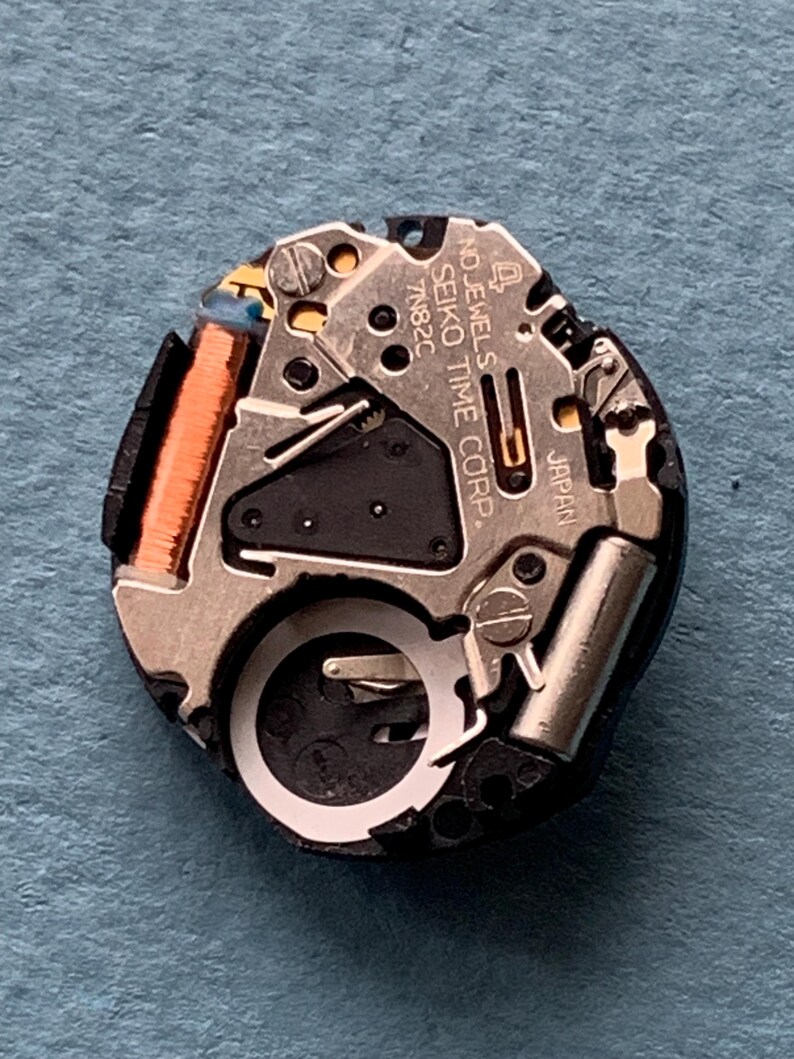 K&ouml;nnte beinhalten: Nahaufnahme eines Seiko-Uhrwerks. Der Mechanismus umfasst silberfarbene Metallkomponenten, eine kupferfarbene Spule und den Text "SEIKO TIME CORP" und "JAPAN". Das Uhrwerk befindet sich vor einem blauen Hintergrund.