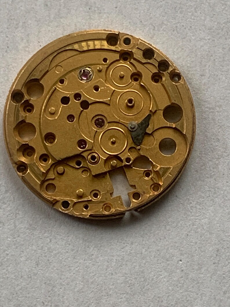 May include: Close-up of a gold-colored watch movement with intricate gears, holes, and small jewels. The circular mechanism has a complex design with various components and a partially cut-out section. The watch part is set against a neutral background.