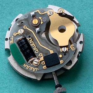 May include: Close-up of a watch movement with the word "ORIENT" printed on the circuit board. The movement has a gold-colored rotor and a green component with the number "32350". The watch mechanism is set against a teal background.