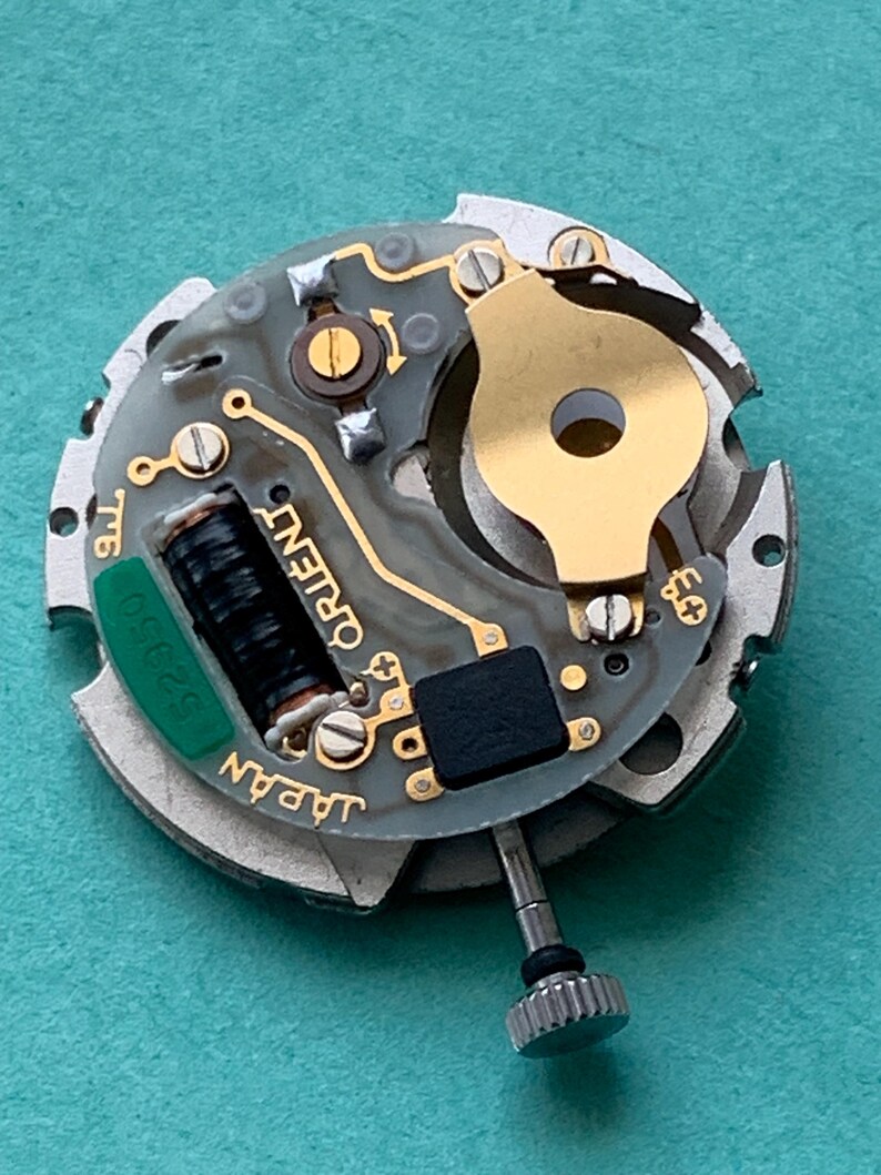May include: Close-up of a watch movement with the word "ORIENT" printed on the circuit board. The movement has a gold-colored rotor and a green component with the number "32350". The watch mechanism is set against a teal background.