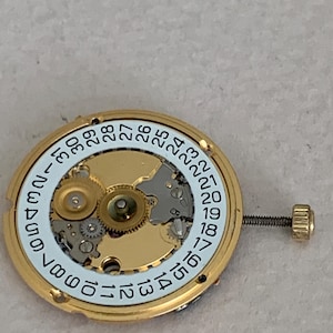 May include: Close-up of a gold-toned watch movement with a white dial featuring black numerals from 1 to 31. The movement has intricate gears and a small screw. The watch component is designed for timekeeping.