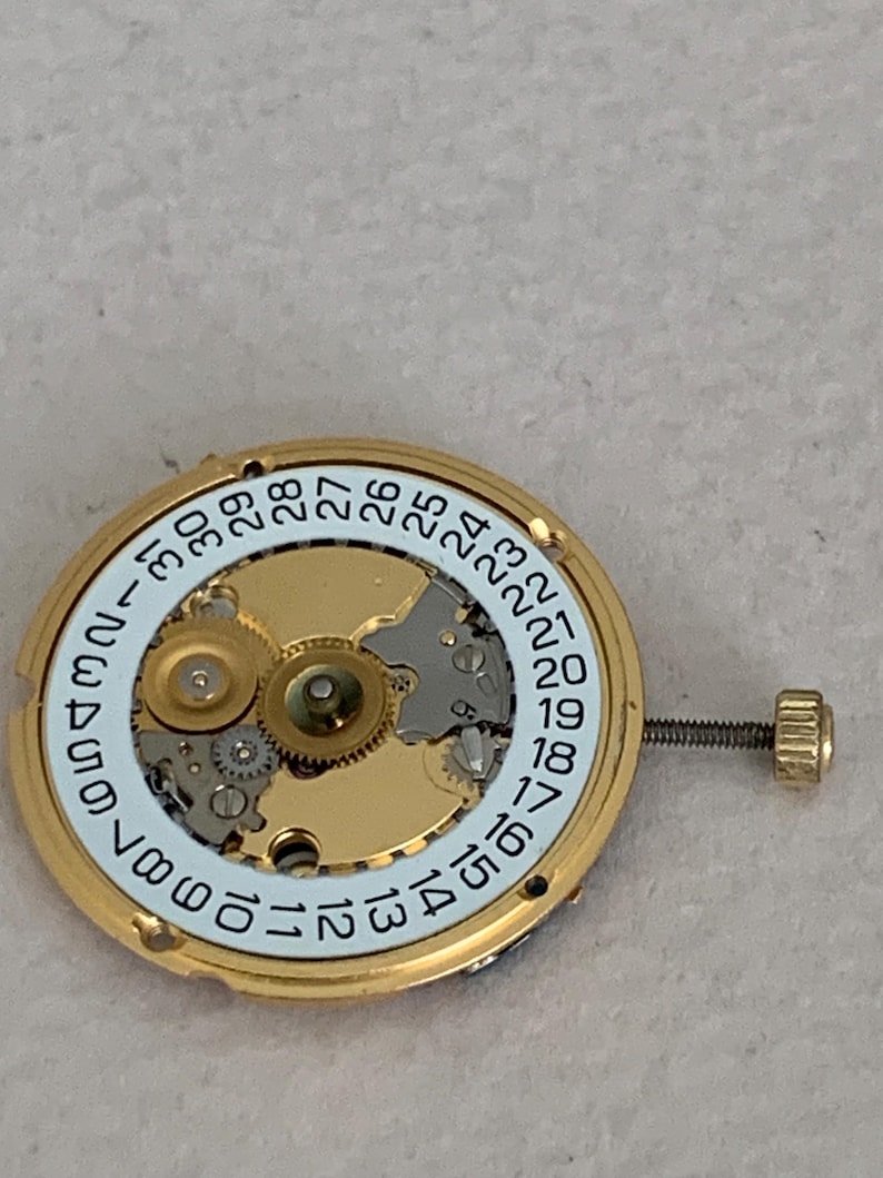 May include: Close-up of a gold-toned watch movement with a white dial featuring black numerals from 1 to 31. The movement has intricate gears and a small screw. The watch component is designed for timekeeping.