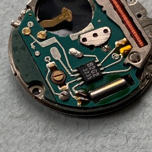 May include: Close-up of a watch movement with a green circuit board. The board features various components, including a small integrated circuit labeled "B202 3235", gold-colored connectors, and a copper coil. The watch mechanism is set against a light gray background.