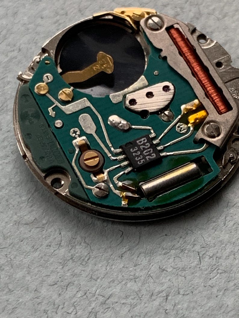 May include: Close-up of a watch movement with a green circuit board. The board features various components, including a small integrated circuit labeled "B202 3235", gold-colored connectors, and a copper coil. The watch mechanism is set against a light gray background.
