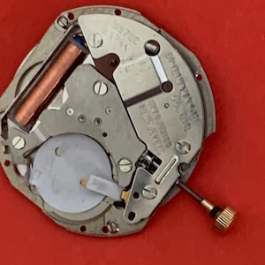 K&ouml;nnte beinhalten: Nahaufnahme eines Uhrwerks mit einem silberfarbenen Metallrahmen. Das Uhrwerk hat eine kupferfarbene Spule, eine wei&szlig;e Kunststoffkomponente und eine goldfarbene Schraube. Der Text "JAPAN" ist sichtbar.
