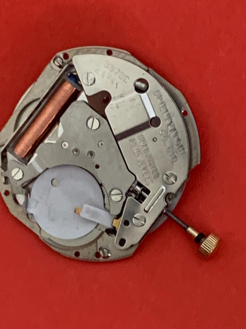 K&ouml;nnte beinhalten: Nahaufnahme eines Uhrwerks mit einem silberfarbenen Metallrahmen. Das Uhrwerk hat eine kupferfarbene Spule, eine wei&szlig;e Kunststoffkomponente und eine goldfarbene Schraube. Der Text "JAPAN" ist sichtbar.