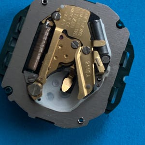 May include: A detailed view of a watch movement, featuring gold-toned gears and mechanisms within a grey metal frame. The base is green plastic. The text "CITIZEN WATCH CO. JAPAN" is visible.