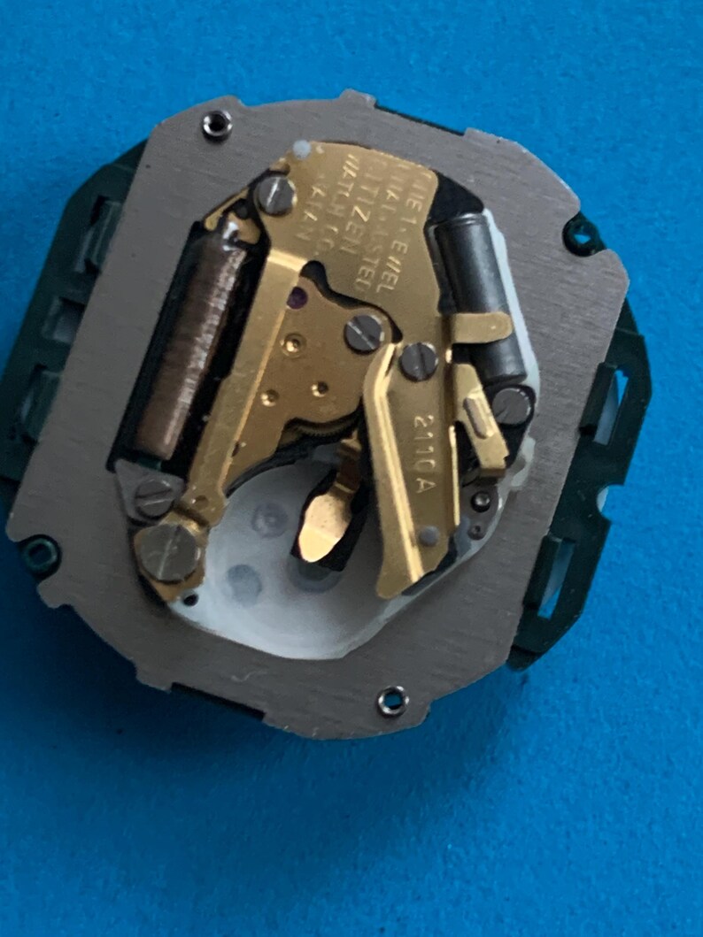 May include: A detailed view of a watch movement, featuring gold-toned gears and mechanisms within a grey metal frame. The base is green plastic. The text "CITIZEN WATCH CO. JAPAN" is visible.