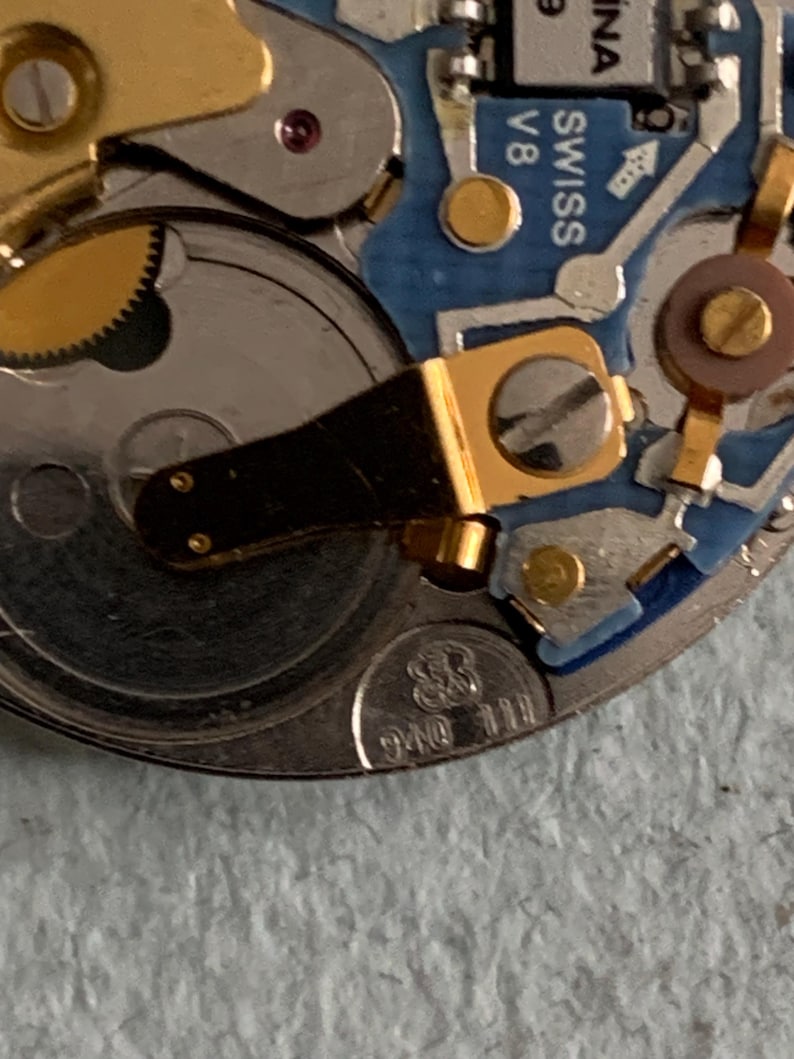 Może przedstawiać: Zbliżenie mechanizmu zegarka z misternymi kołami zębatymi, śrubami i elementami elektronicznymi. Mechanizm zegarka zawiera złote i srebrne metalowe części, niebieską płytkę obwodu drukowanego oraz napis "SWISS V8".