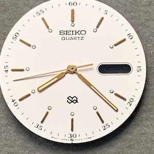 Puede incluir: Una esfera de reloj redonda de color blanco con marcadores de hora y manecillas en tono dorado. La esfera muestra el texto SEIKO QUARTZ, una ventana de fecha en la posición de las tres y el logotipo SQ. Puntos luminosos verdes junto a cada hora.