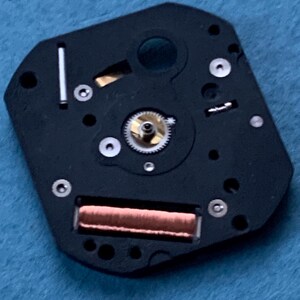 May include: Close-up of a watch movement component. The black, irregularly shaped piece features a central gear, a copper-colored coil, and several small screws. A silver-colored metal bar is also visible. The background is a light blue fabric.
