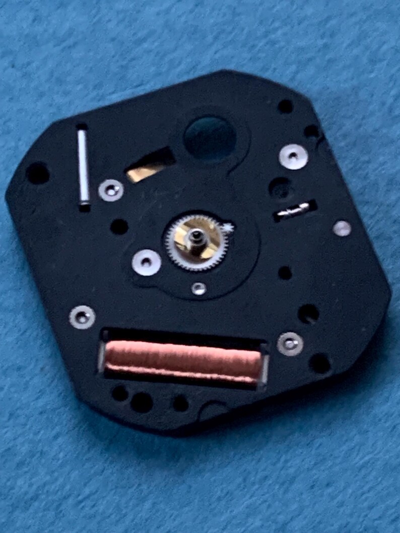 May include: Close-up of a watch movement component. The black, irregularly shaped piece features a central gear, a copper-colored coil, and several small screws. A silver-colored metal bar is also visible. The background is a light blue fabric.