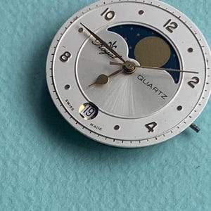 Op de afbeelding: Een zilverkleurige wijzerplaat met gouden wijzers en een maanfase-indicator. De wijzerplaat is voorzien van de woorden "Royal" en "Quartz". De datum is ingesteld op de 19e. De wijzerplaat is gemarkeerd met "Swiss Made".
