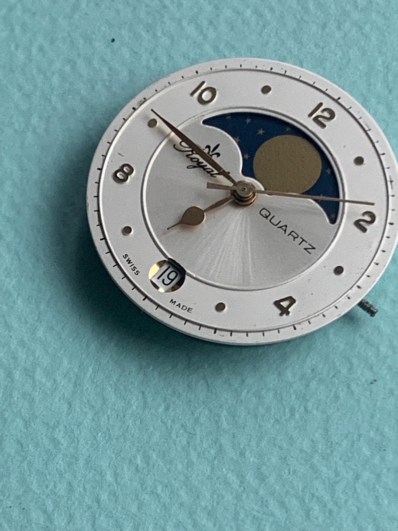Op de afbeelding: Een zilverkleurige wijzerplaat met gouden wijzers en een maanfase-indicator. De wijzerplaat is voorzien van de woorden "Royal" en "Quartz". De datum is ingesteld op de 19e. De wijzerplaat is gemarkeerd met "Swiss Made".