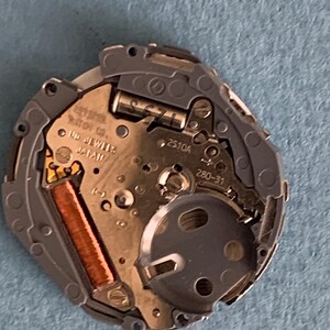 May include: Close-up of a watch movement with intricate gears and components. The watch mechanism features a copper-colored coil, silver-toned metal parts, and the text "MD JEWELS JAPAN". The watch movement is set against a blue background.