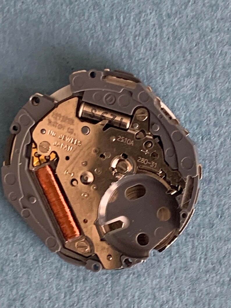 May include: Close-up of a watch movement with intricate gears and components. The watch mechanism features a copper-colored coil, silver-toned metal parts, and the text "MD JEWELS JAPAN". The watch movement is set against a blue background.