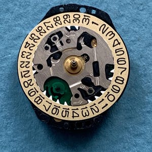 K&ouml;nnte beinhalten: Nahaufnahme eines Uhrwerks mit einem goldfarbenen Datumsring und einem schwarzen Au&szlig;enring. Das Uhrwerk verf&uuml;gt &uuml;ber komplizierte Zahnr&auml;der, einen zentralen Messingstift und eine gr&uuml;ne Komponente. Der Datumsring zeigt die Zahlen 1 bis 31.