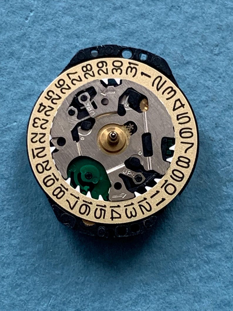 K&ouml;nnte beinhalten: Nahaufnahme eines Uhrwerks mit einem goldfarbenen Datumsring und einem schwarzen Au&szlig;enring. Das Uhrwerk verf&uuml;gt &uuml;ber komplizierte Zahnr&auml;der, einen zentralen Messingstift und eine gr&uuml;ne Komponente. Der Datumsring zeigt die Zahlen 1 bis 31.