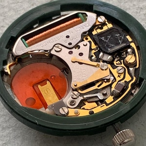 May include: Close-up of a Citizen watch movement, model 7302A, with a green casing. The internal components include a copper coil, gold-colored gears, and a red battery. A silver crown is visible on the side.