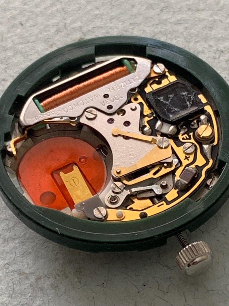 May include: Close-up of a Citizen watch movement, model 7302A, with a green casing. The internal components include a copper coil, gold-colored gears, and a red battery. A silver crown is visible on the side.