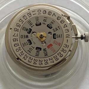 May include: A watch movement with a silver-colored dial and a day-date display. The dial features numbers, days of the week, and a small gold-colored gear. The date is indicated by a red marker. The movement is encased in a clear plastic holder.