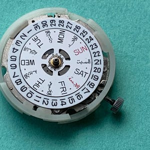 May include: Close-up of a watch movement with a white dial featuring Arabic and English text for days of the week and dates. The dial is surrounded by a white plastic casing and a small metal screw.