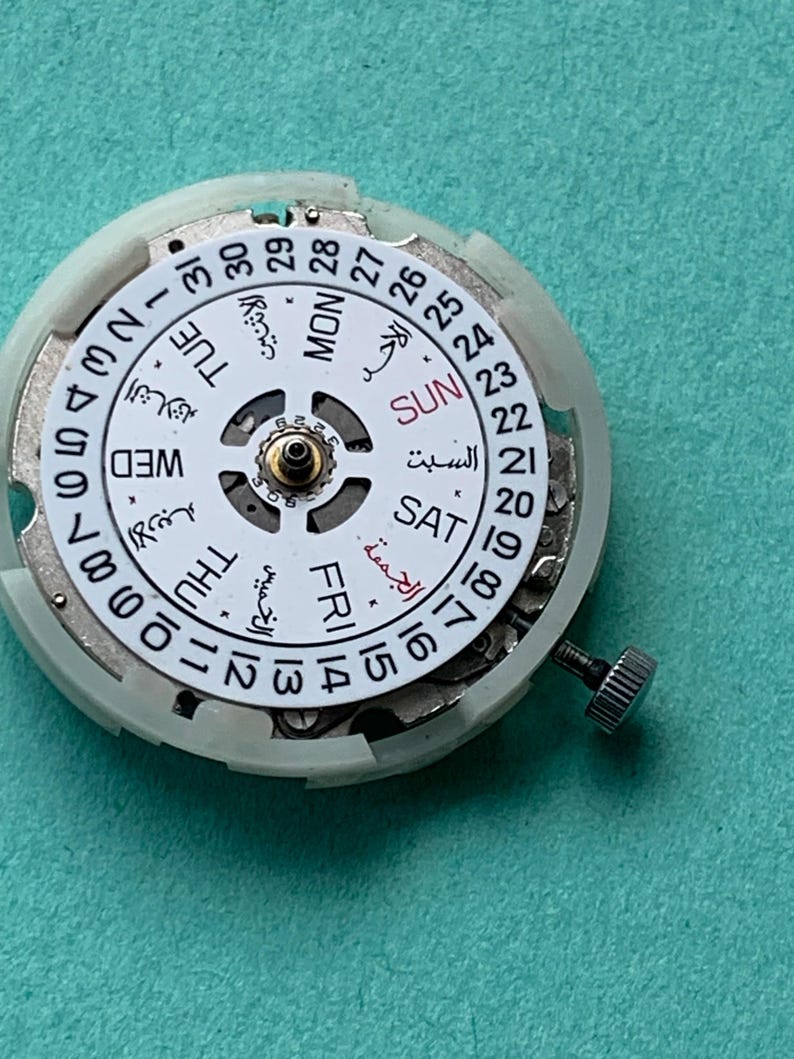 May include: Close-up of a watch movement with a white dial featuring Arabic and English text for days of the week and dates. The dial is surrounded by a white plastic casing and a small metal screw.