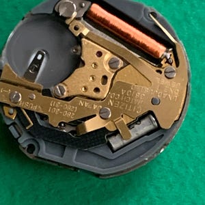 May include: Close-up of a Citizen watch movement. The watch mechanism features gold-toned gears, a copper-colored coil, and the text "Citizen Watch Co. Japan". The watch movement is set against a dark gray background.