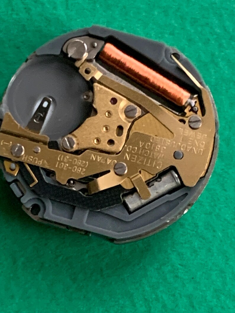 May include: Close-up of a Citizen watch movement. The watch mechanism features gold-toned gears, a copper-colored coil, and the text "Citizen Watch Co. Japan". The watch movement is set against a dark gray background.