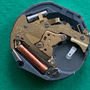 May include: Close-up of a watch movement with gold-colored gears and a copper-colored coil. The watch mechanism is set in a gray casing with the text "CITIZEN JAPAN" visible. A battery is also present.