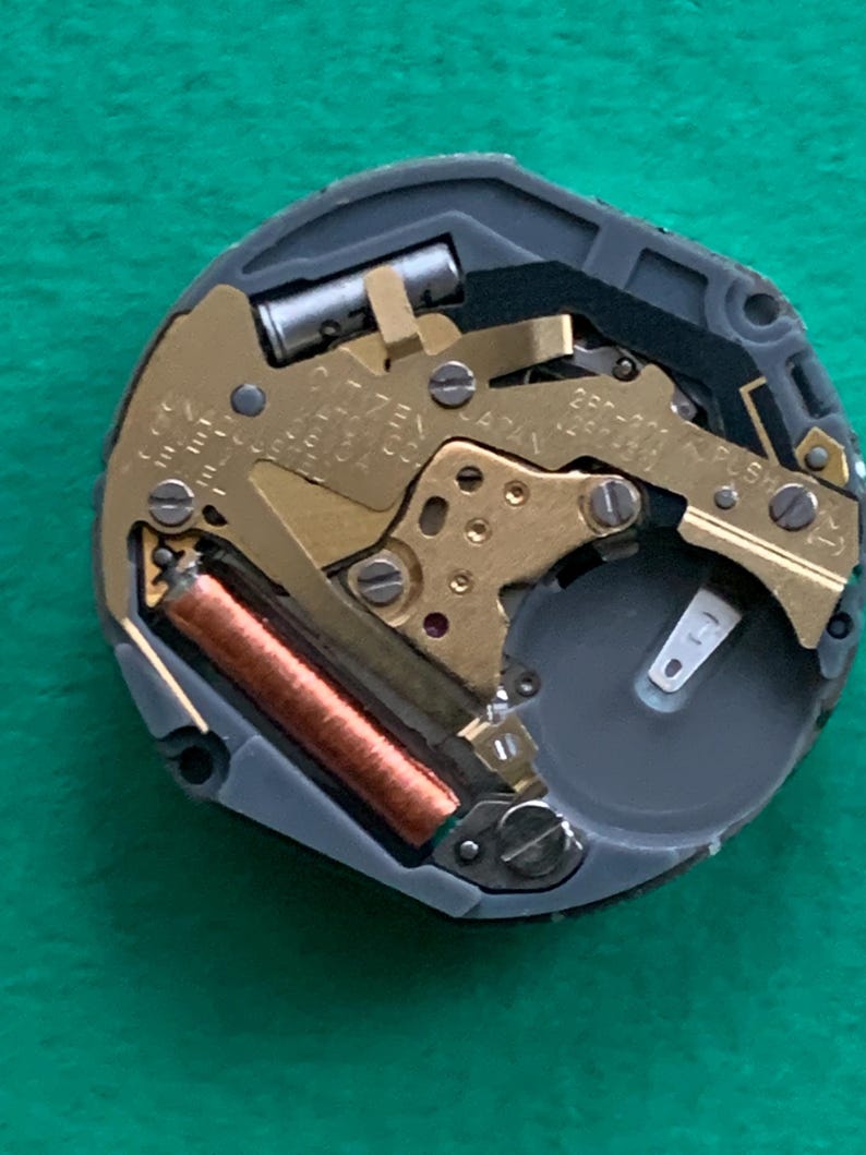 May include: Close-up of a watch movement with gold-colored gears and a copper-colored coil. The watch mechanism is set in a gray casing with the text "CITIZEN JAPAN" visible. A battery is also present.