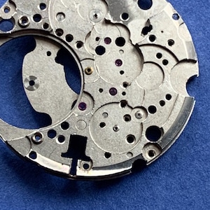Puede incluir: Primer plano de un movimiento de reloj plateado con numerosos agujeros, engranajes y peque&ntilde;os componentes. El intrincado dise&ntilde;o presenta formas circulares y curvas, con algunas joyas moradas. El fondo es azul liso.
