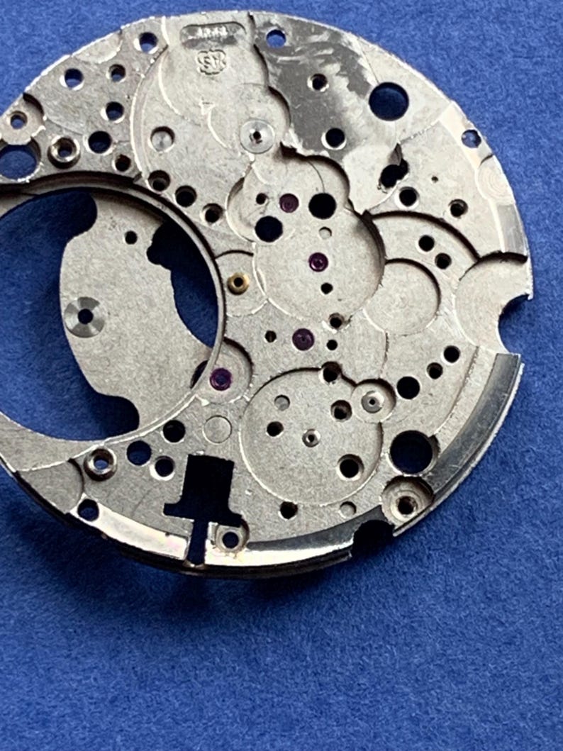 Puede incluir: Primer plano de un movimiento de reloj plateado con numerosos agujeros, engranajes y peque&ntilde;os componentes. El intrincado dise&ntilde;o presenta formas circulares y curvas, con algunas joyas moradas. El fondo es azul liso.