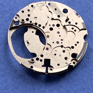 Puede incluir: Primer plano de un movimiento de reloj plateado con intrincados engranajes, agujeros y una gran abertura curva. El mecanismo del reloj tiene un dise&ntilde;o complejo con varios componentes peque&ntilde;os y marcas. El fondo es azul liso.