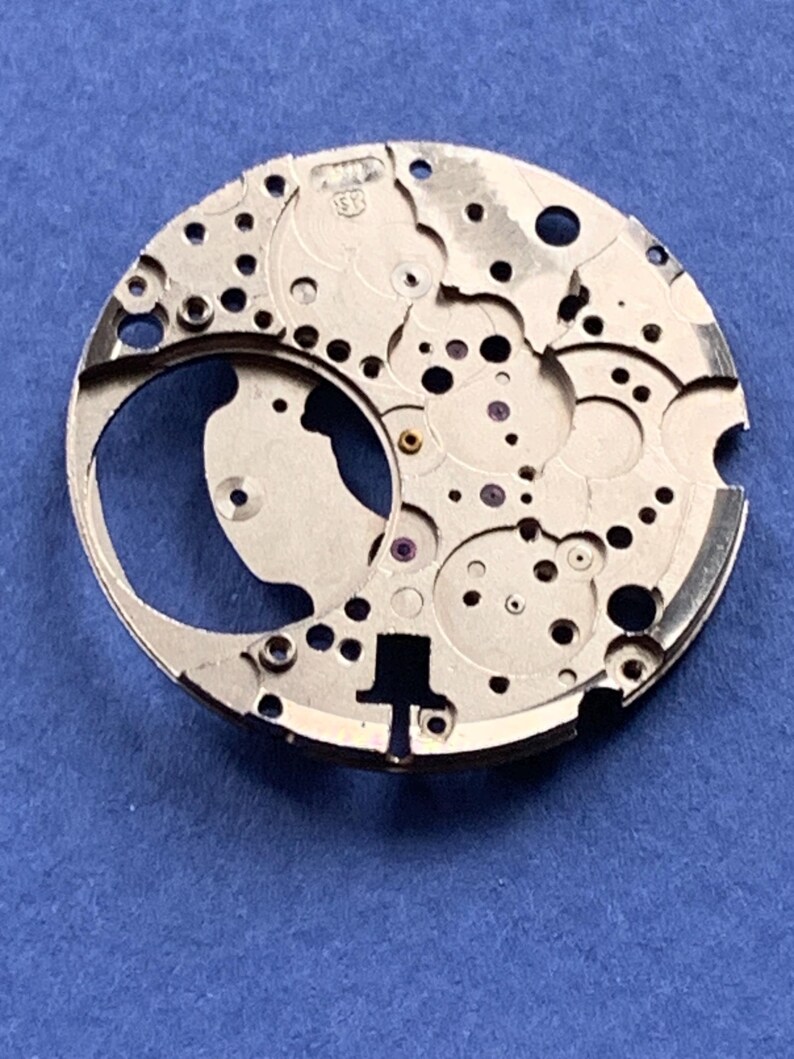 Puede incluir: Primer plano de un movimiento de reloj plateado con intrincados engranajes, agujeros y una gran abertura curva. El mecanismo del reloj tiene un dise&ntilde;o complejo con varios componentes peque&ntilde;os y marcas. El fondo es azul liso.