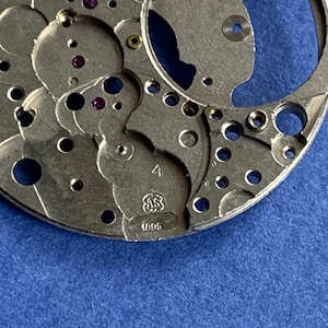 Puede incluir: Primer plano de un movimiento de reloj plateado con intrincados engranajes, ruedas y joyas de rub&iacute;. Los componentes met&aacute;licos est&aacute;n detallados con varios agujeros y formas. El texto "4", "AS" y "1695" es visible.