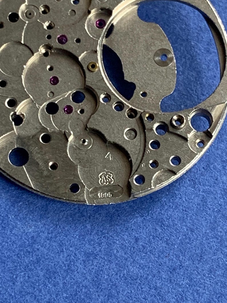 Puede incluir: Primer plano de un movimiento de reloj plateado con intrincados engranajes, ruedas y joyas de rub&iacute;. Los componentes met&aacute;licos est&aacute;n detallados con varios agujeros y formas. El texto "4", "AS" y "1695" es visible.