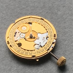 May include: Close-up of a gold-toned watch movement with visible gears, a date dial, and a screw. The dial features numbers from 1 to 31, indicating the days of the month. The intricate mechanical components are clearly displayed.