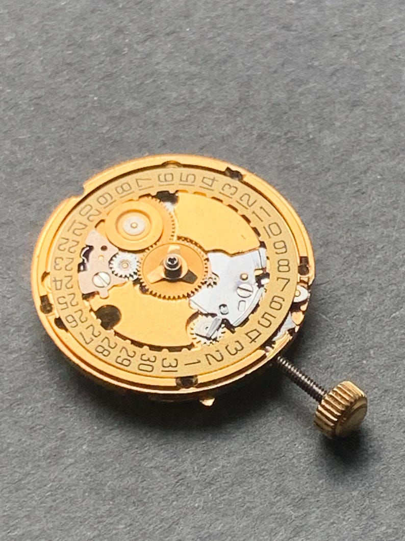 May include: Close-up of a gold-toned watch movement with visible gears, a date dial, and a screw. The dial features numbers from 1 to 31, indicating the days of the month. The intricate mechanical components are clearly displayed.