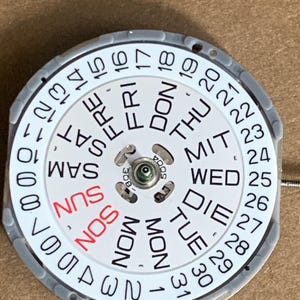 May include: A watch calendar wheel with a white face, featuring days of the week in English and numbers 1-31. The day "SUN" is in red. The wheel is surrounded by a gray ring.